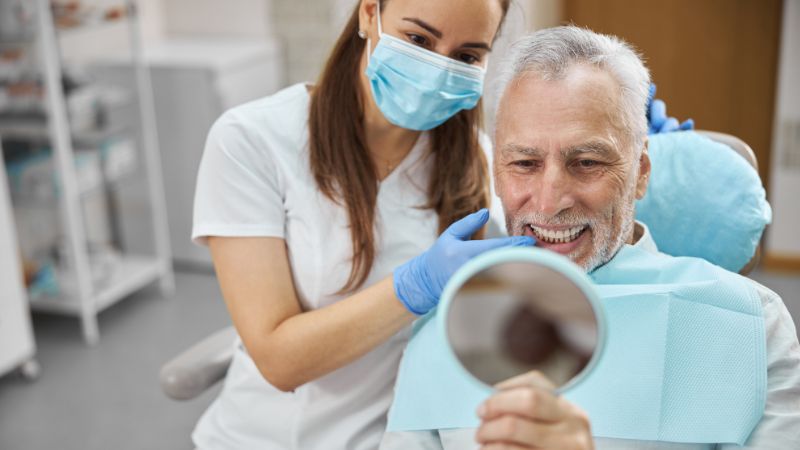 Paziente anziano controlla il risultato di un impianto dentale allo specchio con l’assistenza della dentista: fase post-operatoria di verifica dell’integrazione protesica.