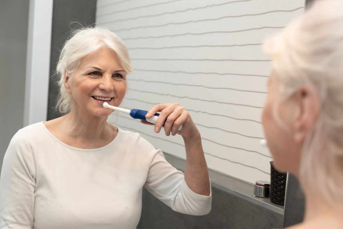 Donna matura utilizza uno spazzolino elettrico davanti allo specchio: corretta igiene orale quotidiana per mantenere puliti gli impianti dentali.