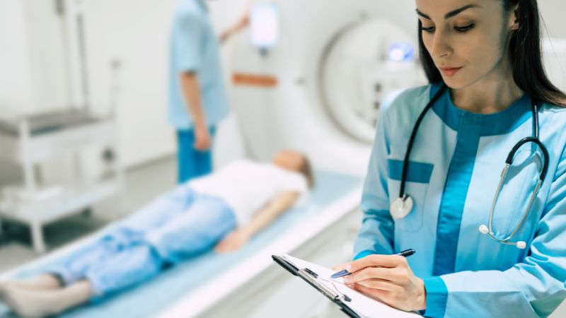 Una dottoressa in primo piano compila una cartella clinica, mentre sullo sfondo un paziente si prepara per una risonanza magnetica. La scena illustra la fase di anamnesi, cruciale per verificare la presenza di impianti dentali prima dell'esame