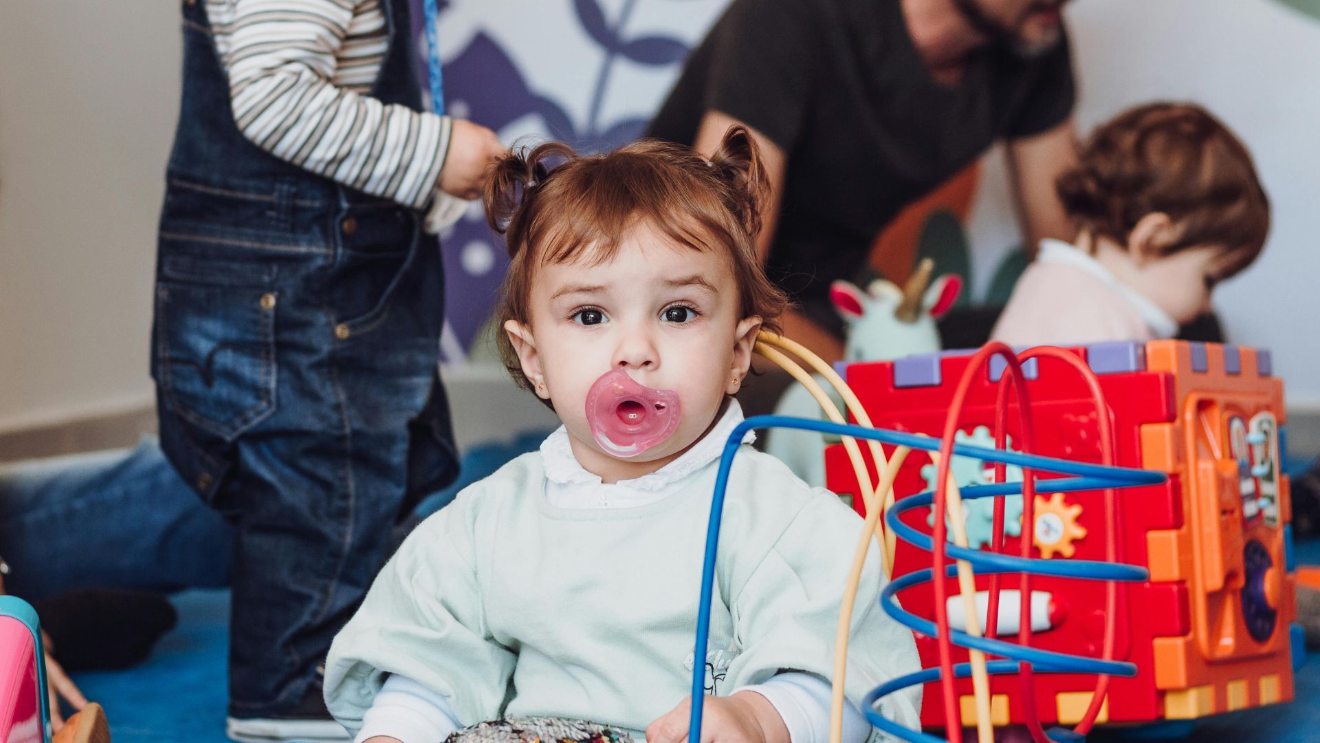In un contesto di gioco, una bambina con un ciuccio rosa in bocca guarda verso l'obiettivo, illustrando l'uso del succhietto, potenziale causa di malocclusioni o denti storti se protratto nel tempo