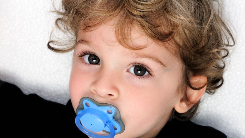 Un bambino piccolo con i capelli ricci e grandi occhi guarda la fotocamera mentre tiene in bocca un ciuccio, a rappresentare una delle abitudini che possono influenzare lo sviluppo della dentatura