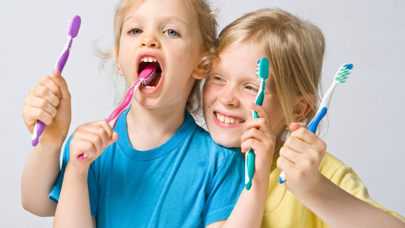 Due bambini rendono il momento dell'igiene orale un gioco: uno si spazzola la lingua con uno spazzolino rosa mentre l'altro sorride, entrambi con i loro spazzolini in mano.