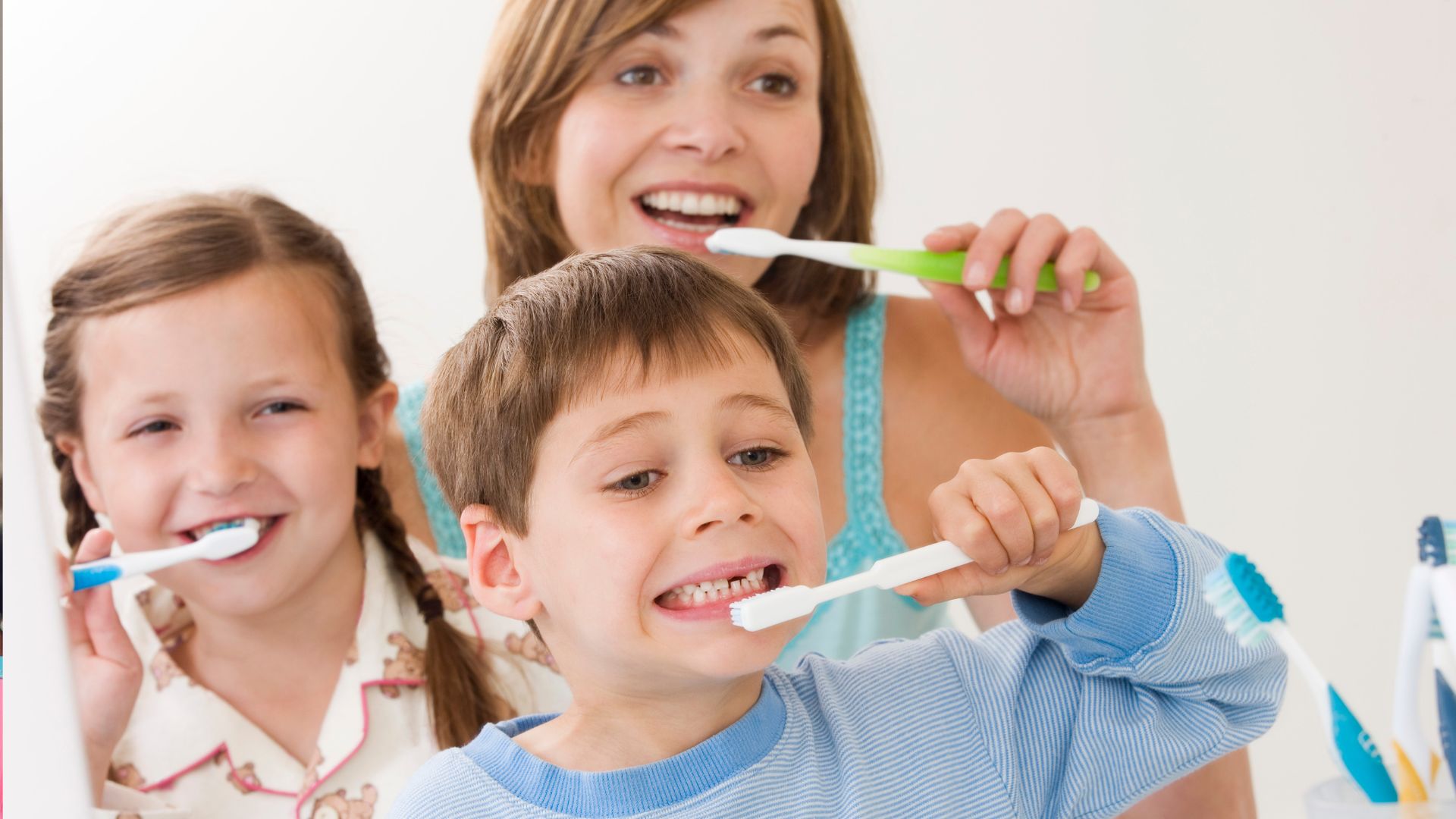 Un adulto dà il buon esempio a due bambini lavando i denti insieme a loro. Il bambino in primo piano si spazzola i denti con attenzione, mostrando come la routine familiare aiuti a imparare la corretta igiene orale